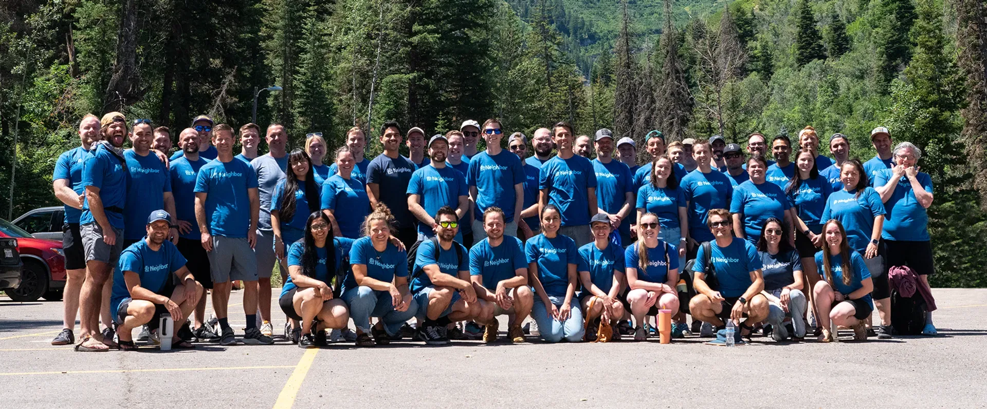 Large diverse team of employees in blue company shirts standing together for a group photo with mountains and forest in the background