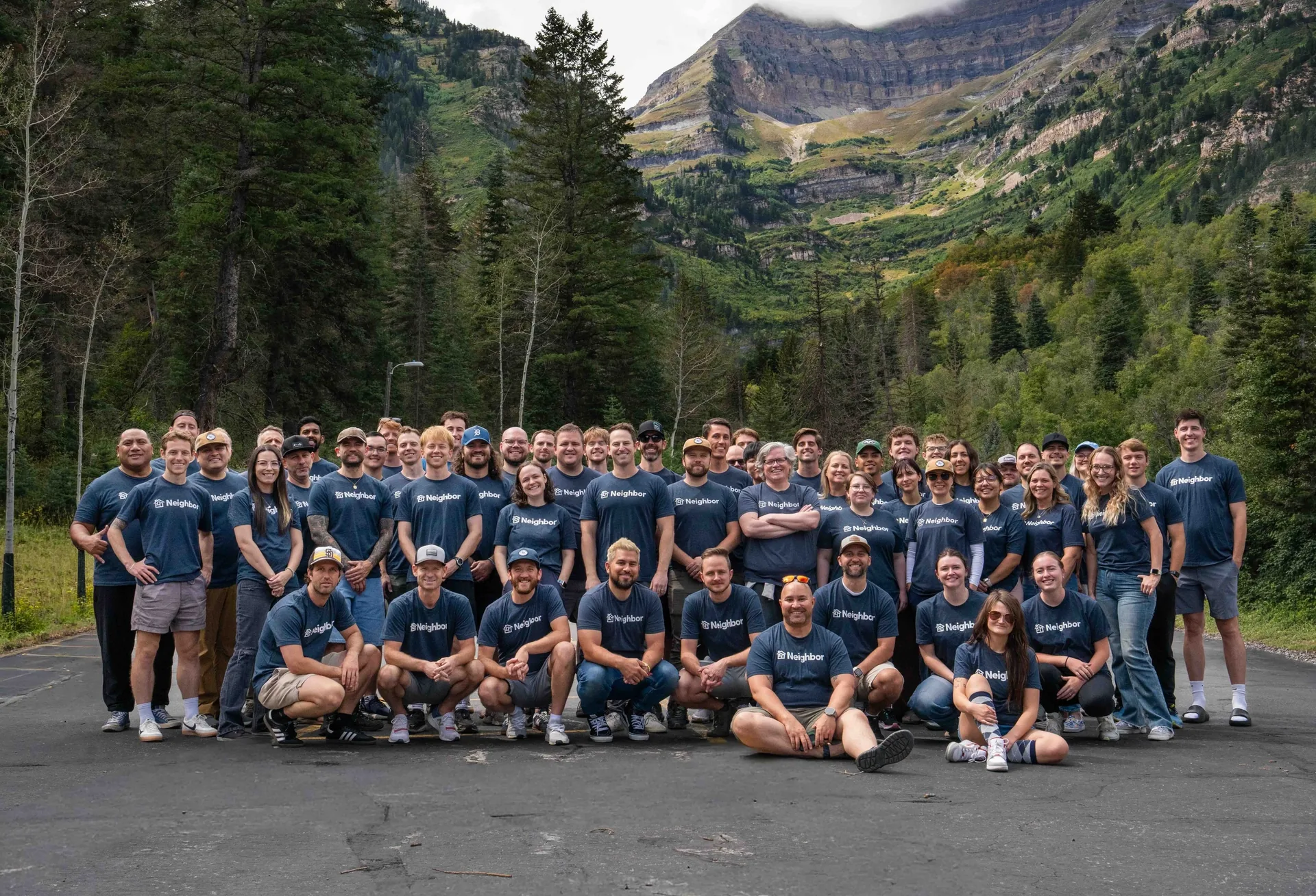 Large company team photo of approximately 60 people in matching dark blue Neighbor branded shirts standing and crouching on an asphalt road in a forested mountain valley with snow-capped peaks visible in the background.